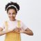 Love my fans. Portrait of cute carefree and confident young female blogger with dark skin in yellow dungarees and headband, showing heart sign over chest and smiling with caring and flirty expression.